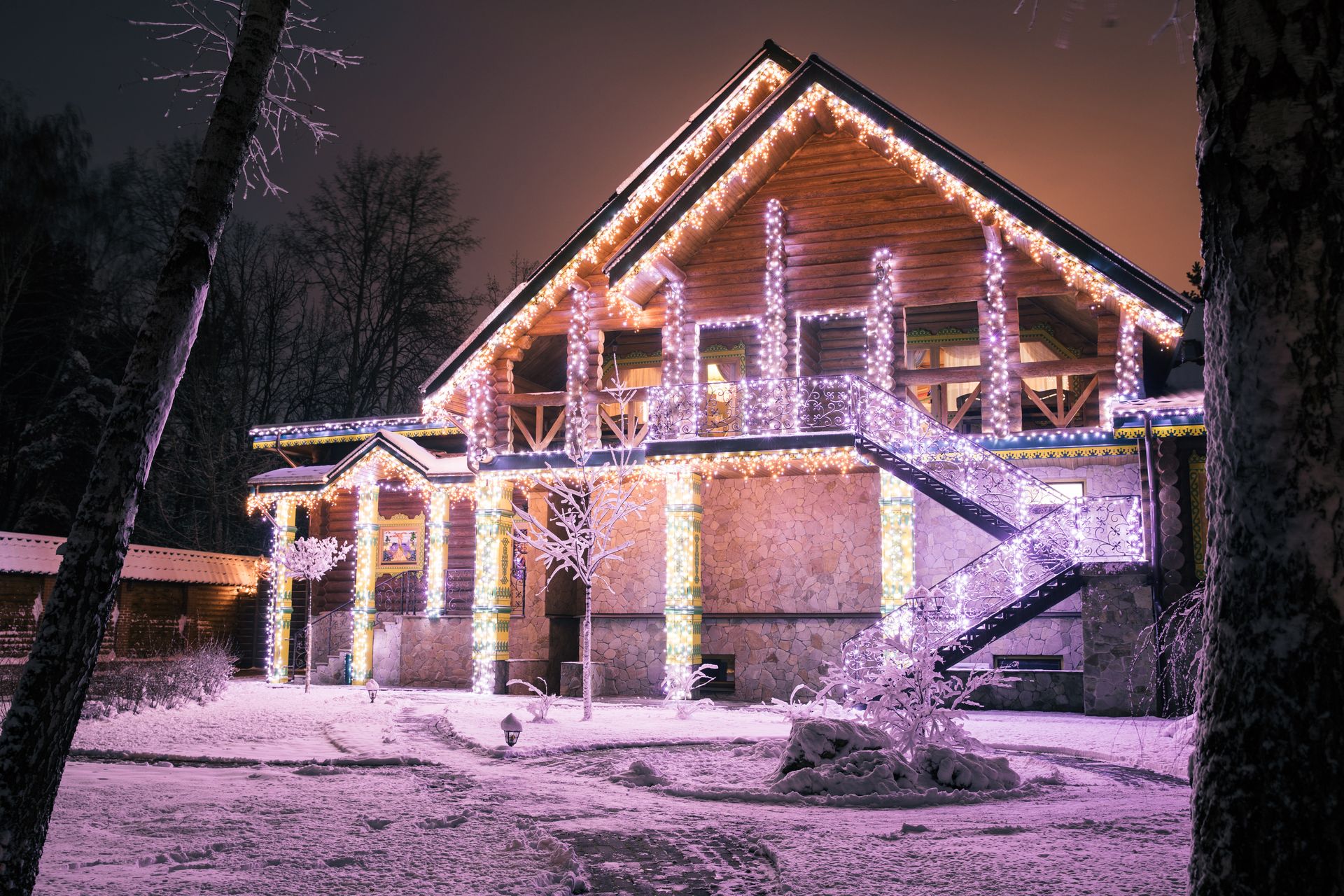 Snow-covered house at night with purple holiday lights and lit windows in a winter scene