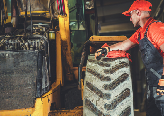 Mechanic in orange shirt and hat wipes a large tractor tire. Dark workshop setting.