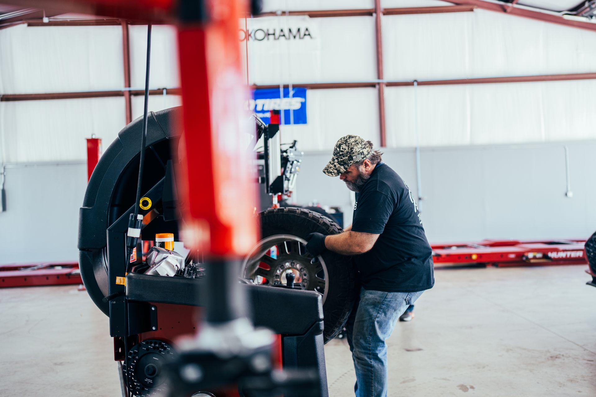 Mechanic balancing a tire in a garage, wearing a hat, black shirt, and jeans.