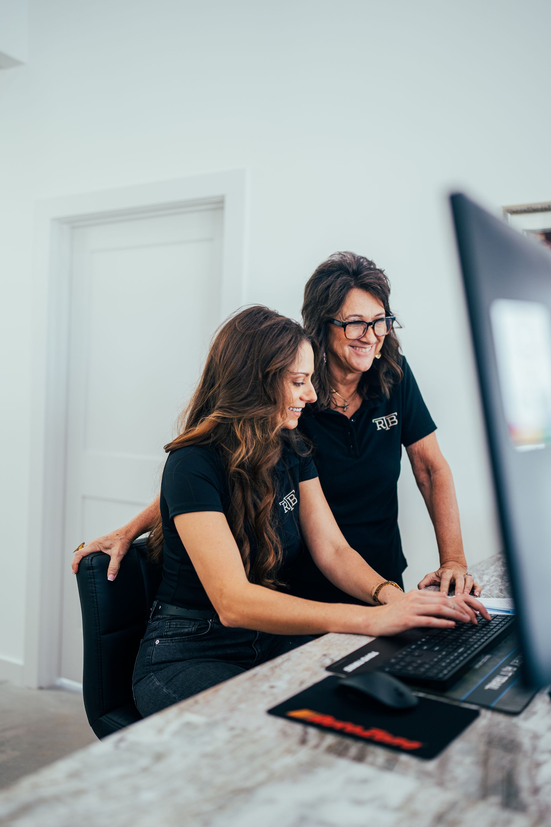 Two people looking at a computer monitor. One types, the other leans over her shoulder, both wear black shirts.
