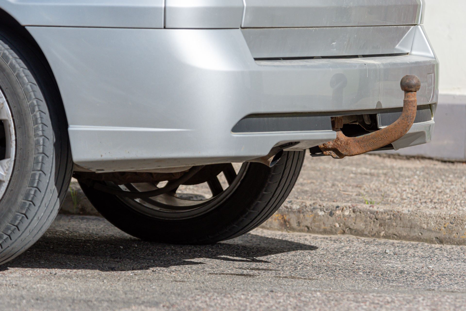 Silver car with a rusty tow hitch parked near a curb, viewed from the rear.