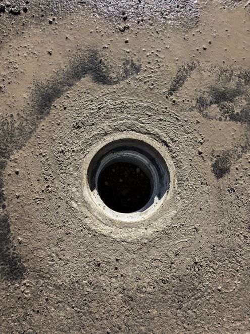 Close-up of a circular black drain opening set in rough beige concrete or asphalt.