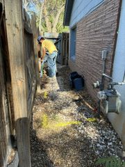 Worker clearing debris in a narrow side yard beside a brick house and wooden fence.