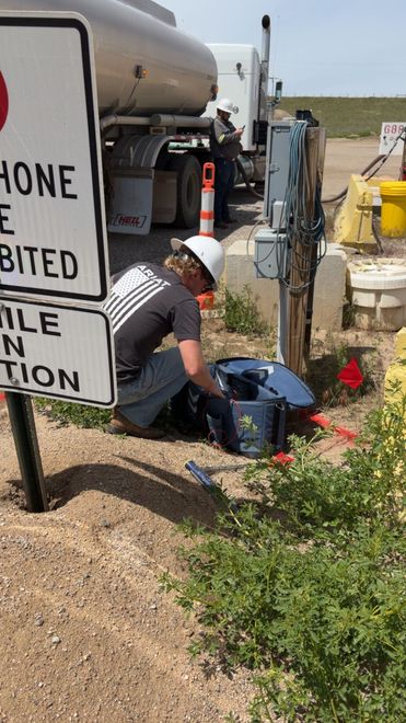 Worker kneeling beside utility truck and equipment at roadside worksite with construction sign