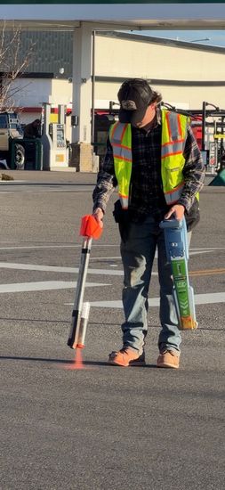 Worker in a reflective vest using a surveying rod on a paved worksite.