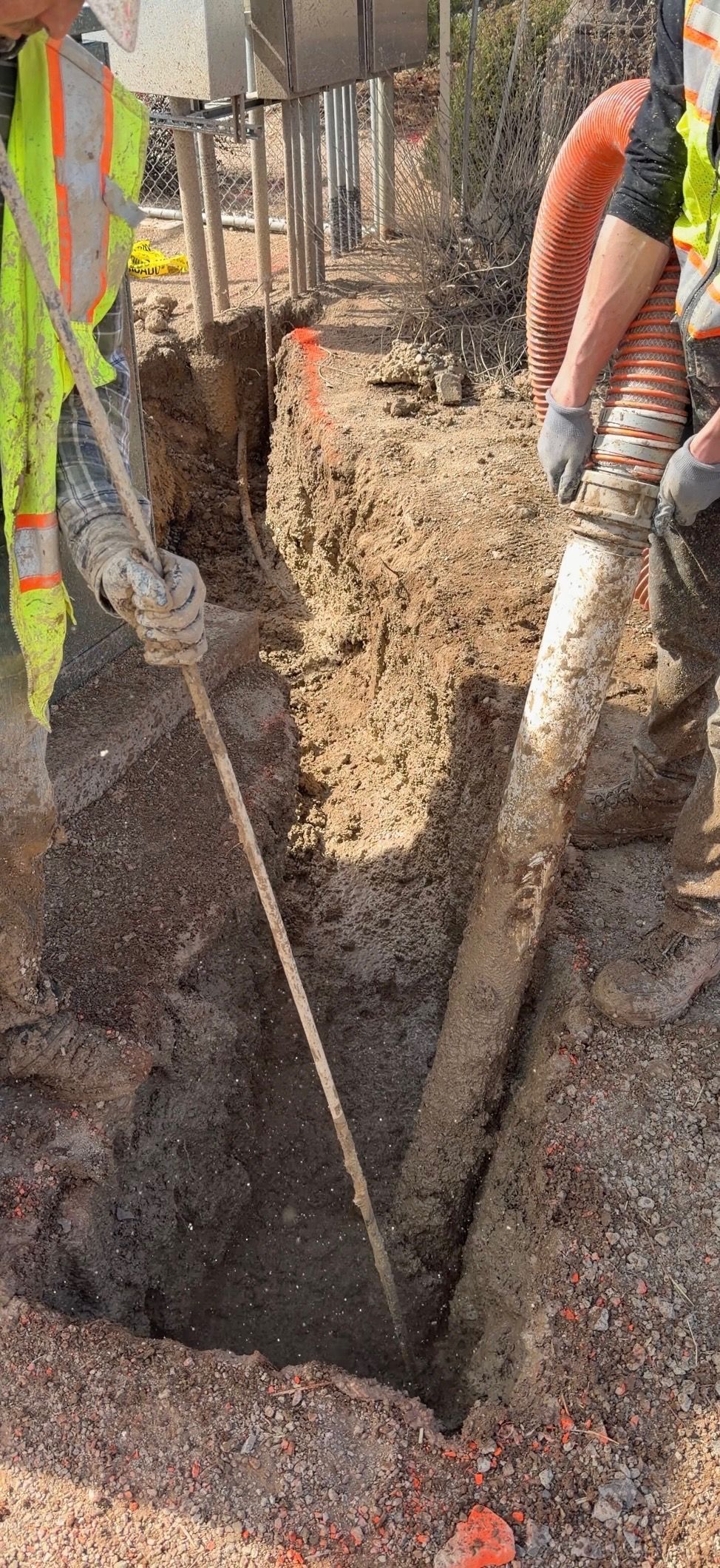 Workers in safety vests dig a deep trench beside a tree-lined dirt road.