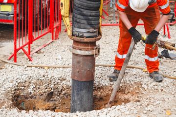 Construction worker in orange gear drilling into a gravel worksite beside a large borehole.