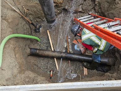 Worker in safety gear handling large black pipe in a trench beside an orange ladder and green hose