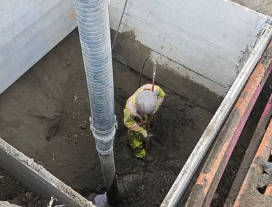 Worker in yellow safety gear kneeling in a muddy concrete pit beside a large pipe
