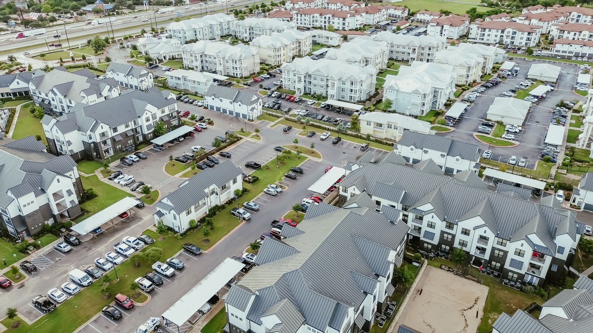 Aerial view of a large apartment complex with gray roofs, parking lots, and green landscaping.