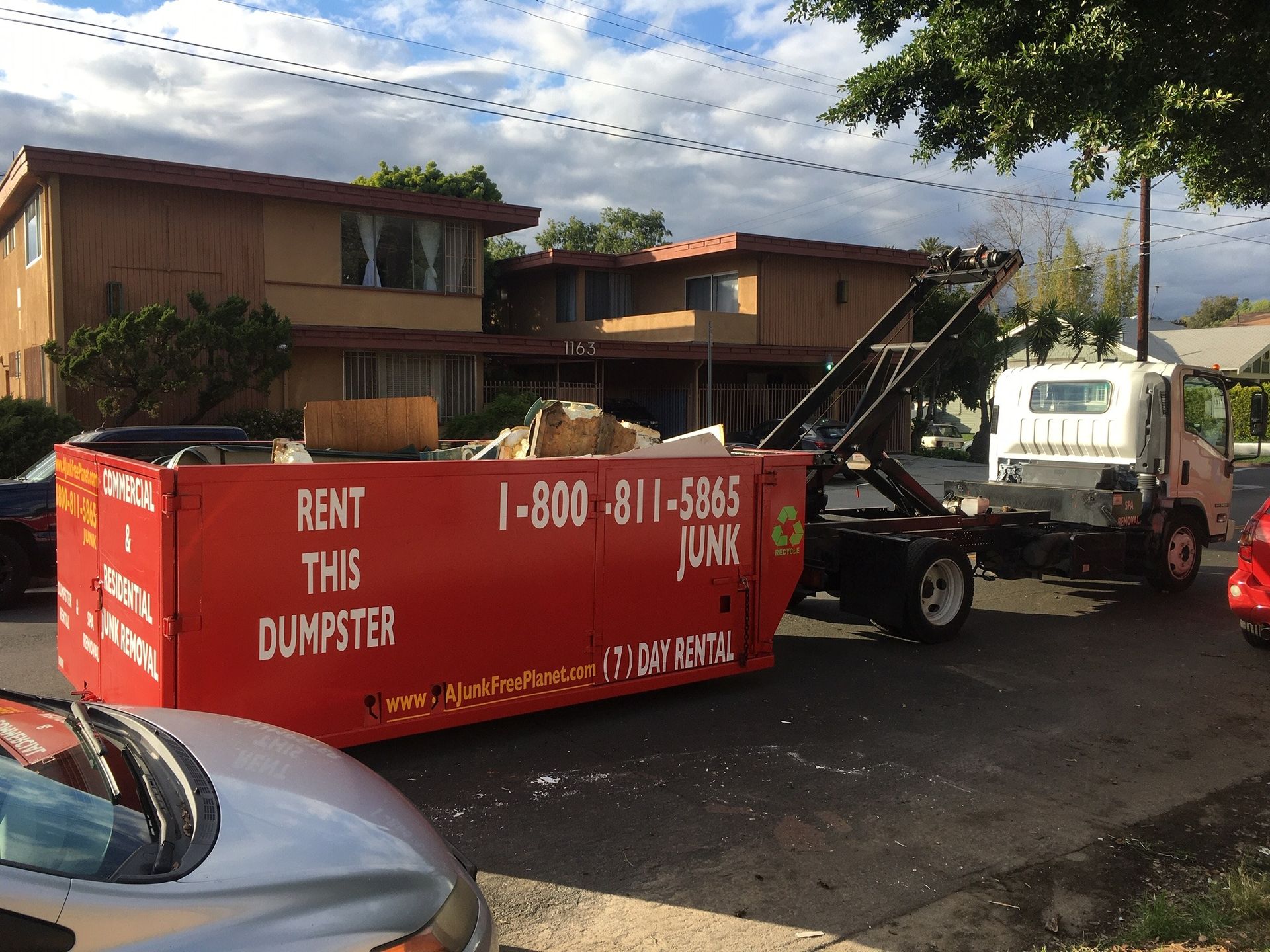 Red dumpster being loaded by truck; apartments in the background. Junk removal service.