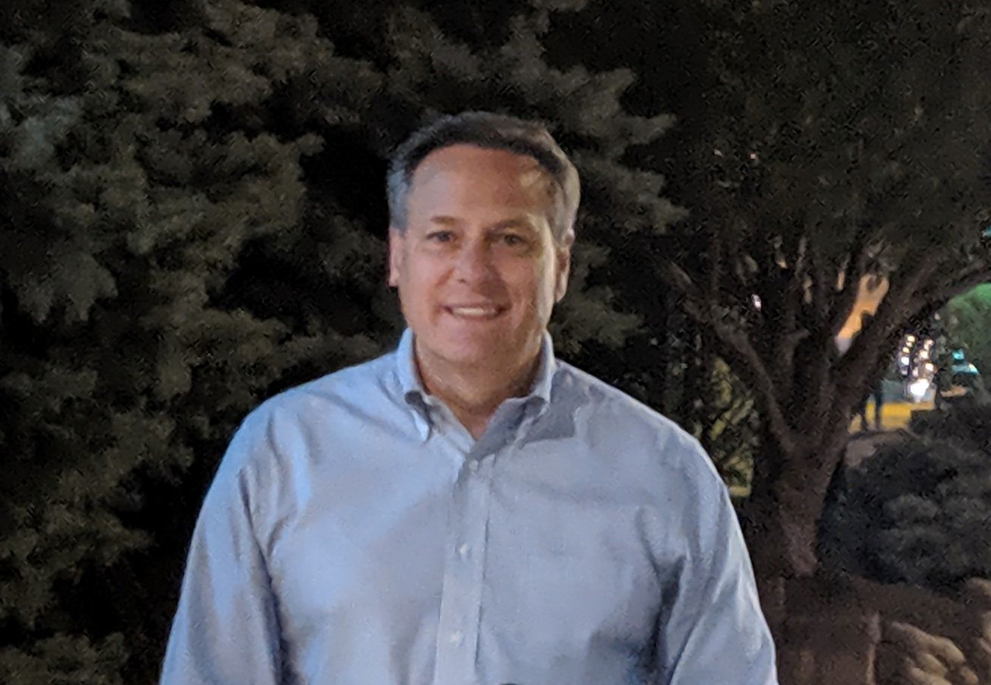 Smiling man in a light blue shirt poses outdoors at dusk with trees in the background.