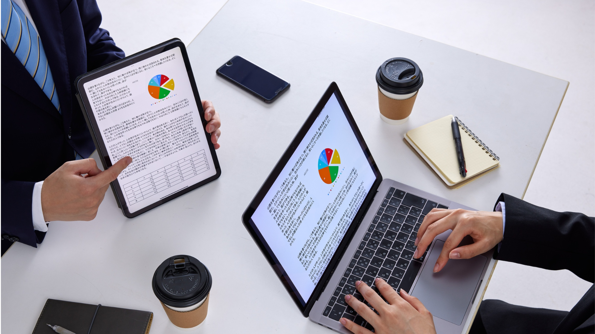 Two people reviewing charts on tablet and laptop at a white table with coffee cups and a notebook