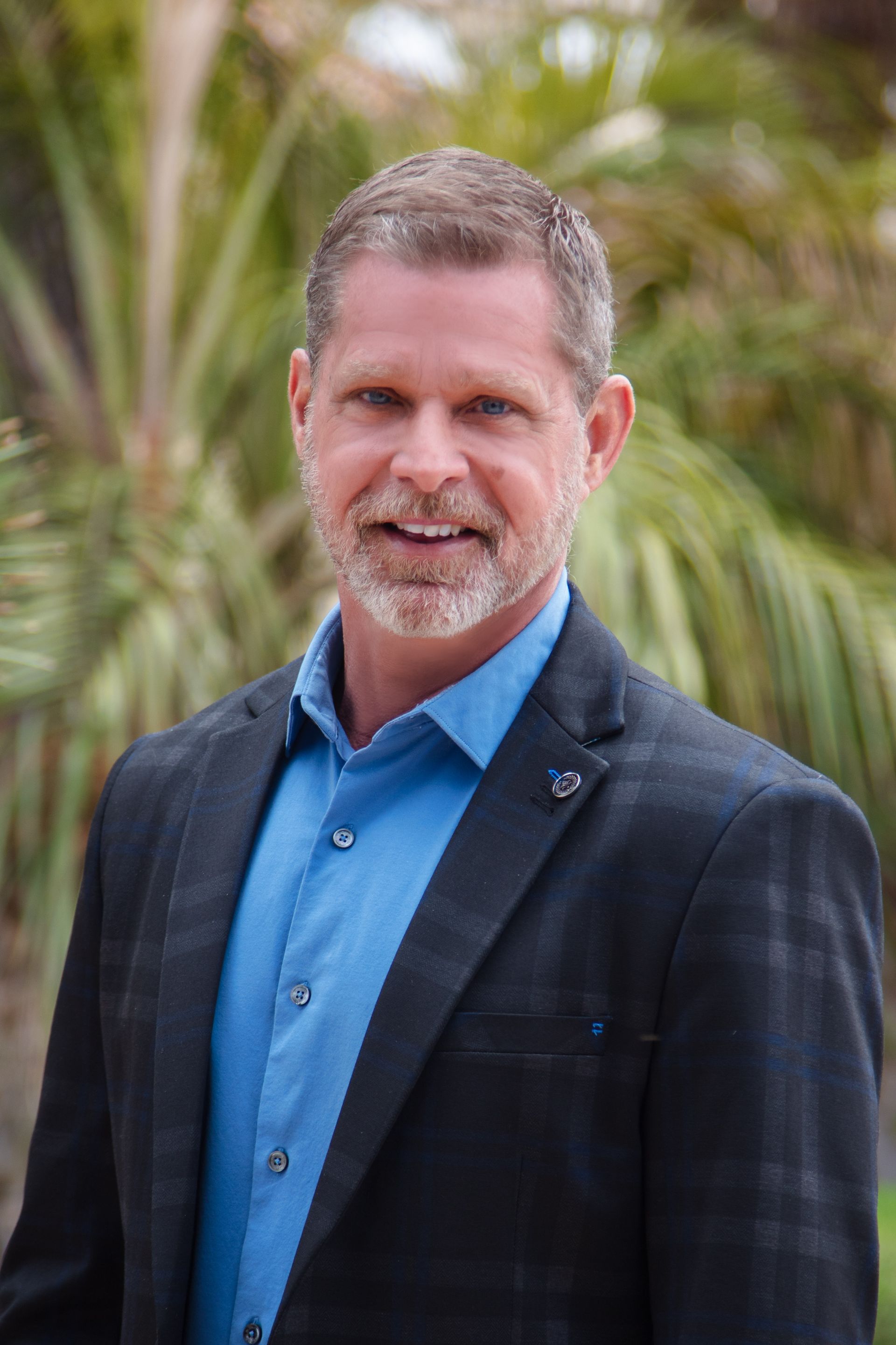 Man in a suit smiling outdoors, blue shirt, gray hair, and beard.