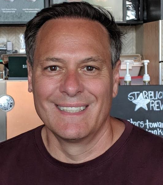 Man smiling in a maroon shirt, in a Starbucks.