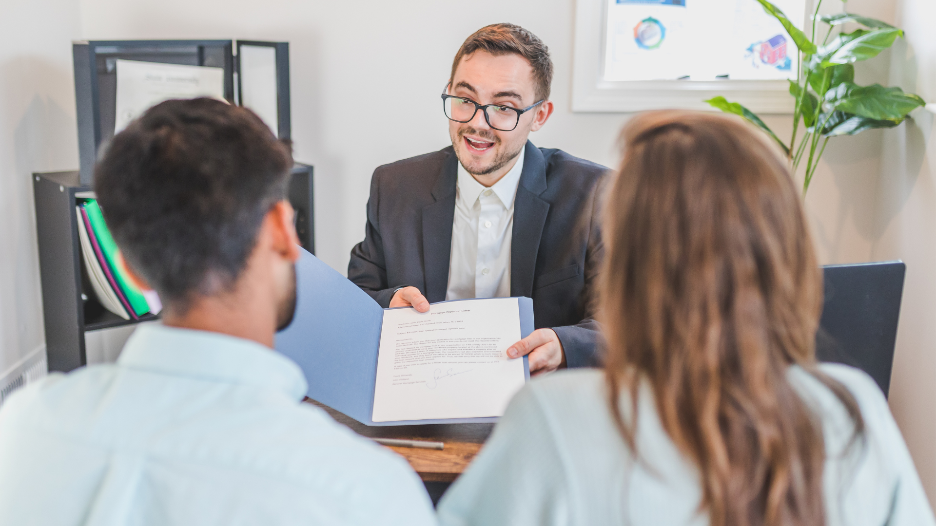 Job interview in an office, with a smiling man in a suit speaking to two seated candidates.