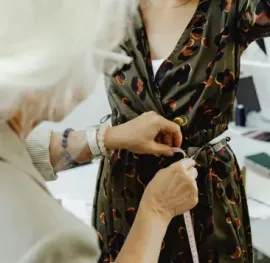 A seamstress measures a dress on a person in a brightly lit room with office supplies on a shelf.