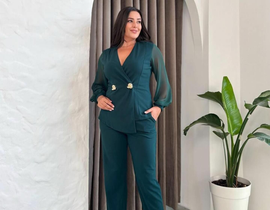 Woman in a green pantsuit with sheer sleeves, smiling, gold heels, standing indoors by a plant.