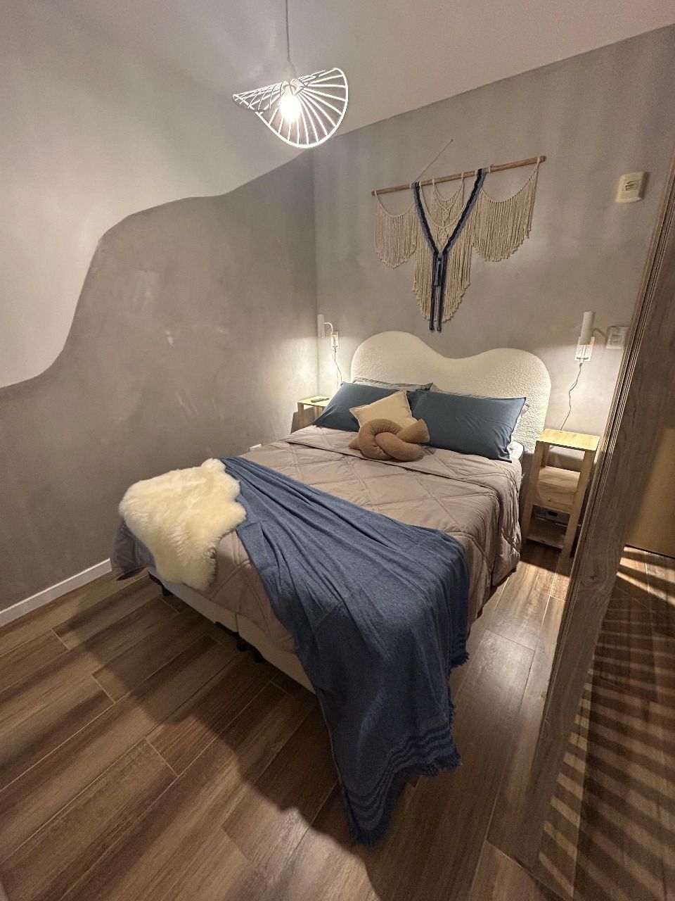 Dormitorio con cama, mesitas de noche y decoración, incluyendo un tapiz de macramé y una manta azul.
