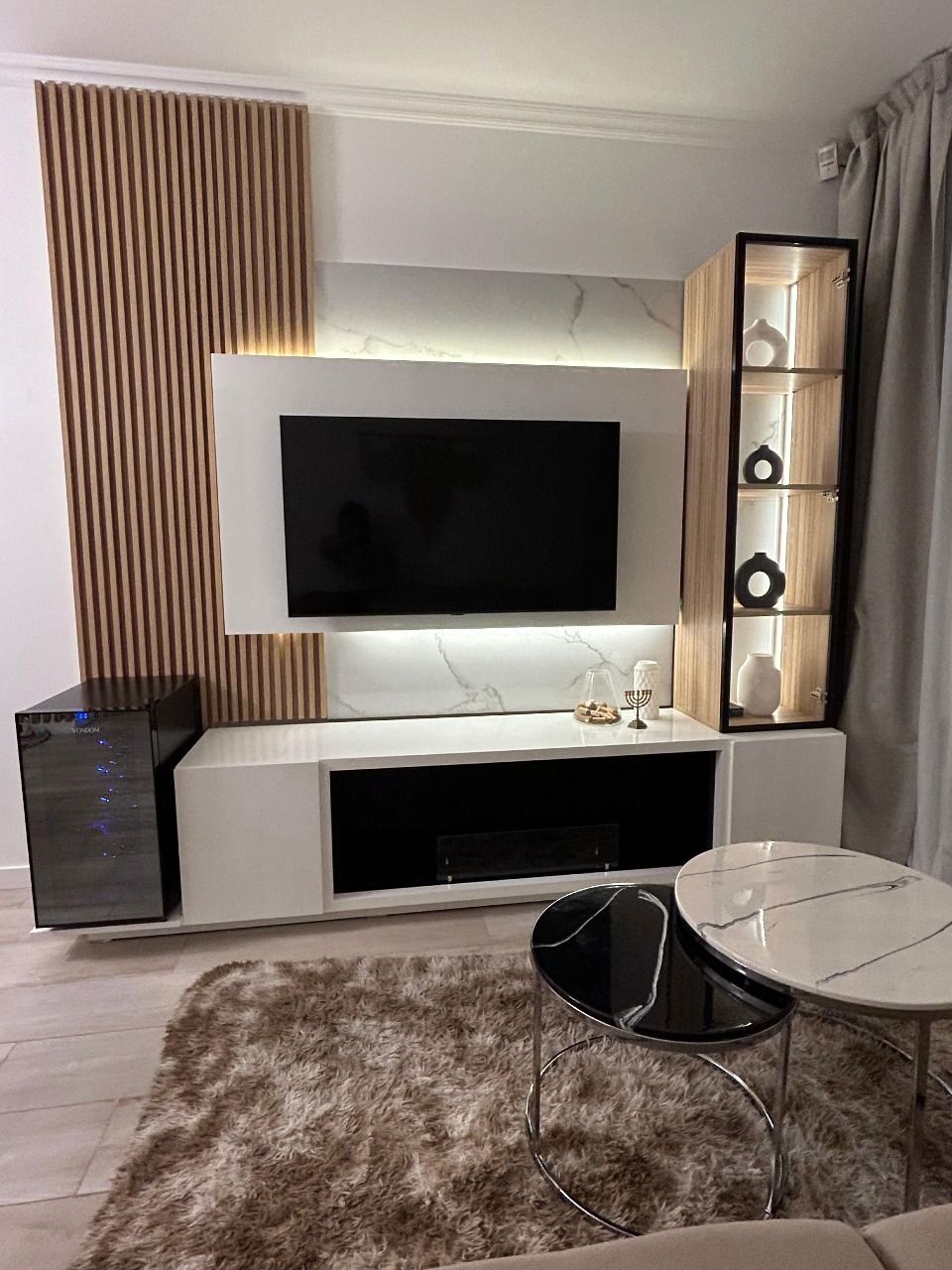 Salón moderno con centro de entretenimiento blanco y panelado en madera, televisor y vitrina.
