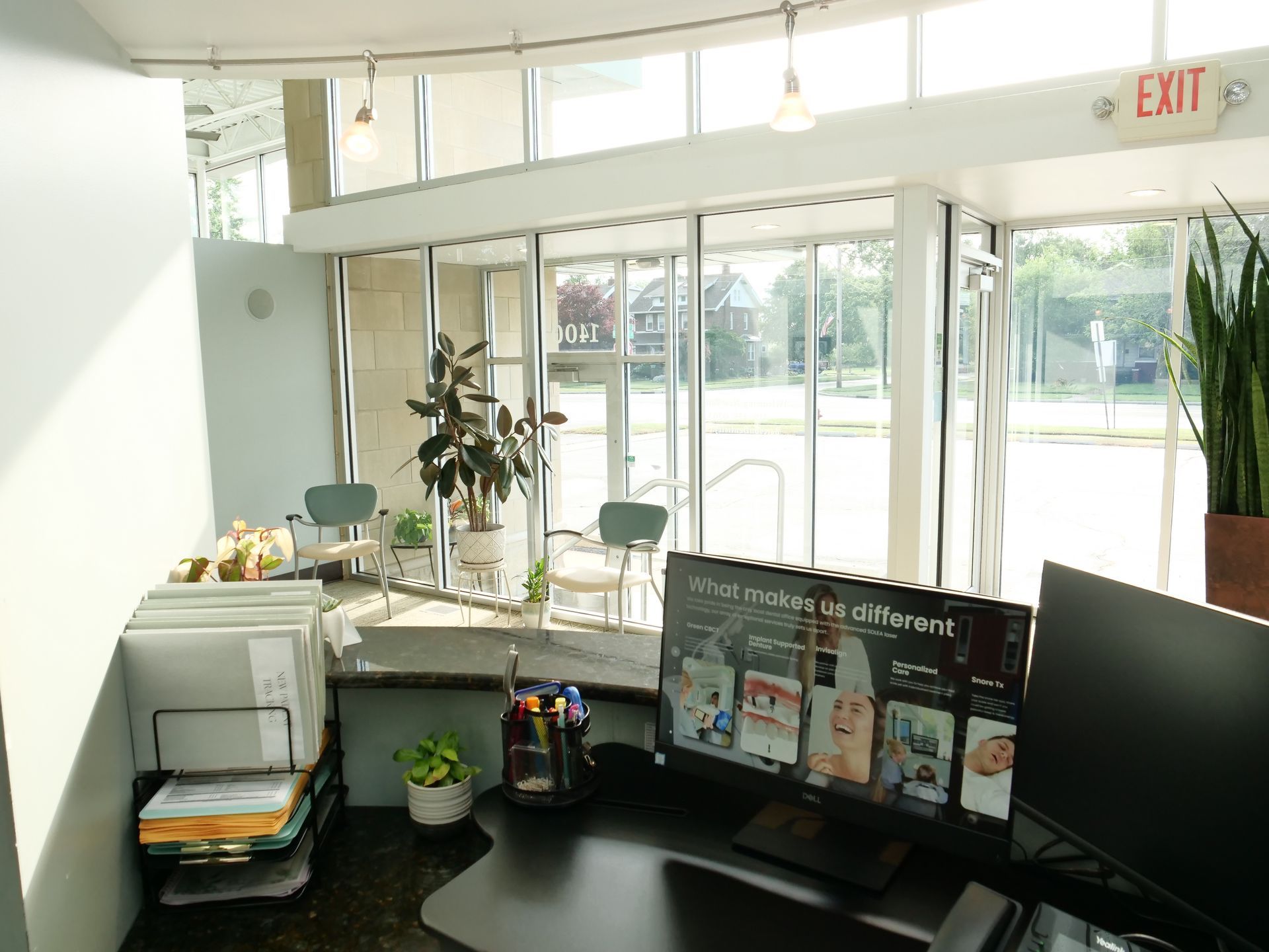 Bright office reception desk with monitors, plants, and sunlight through glass doors.
