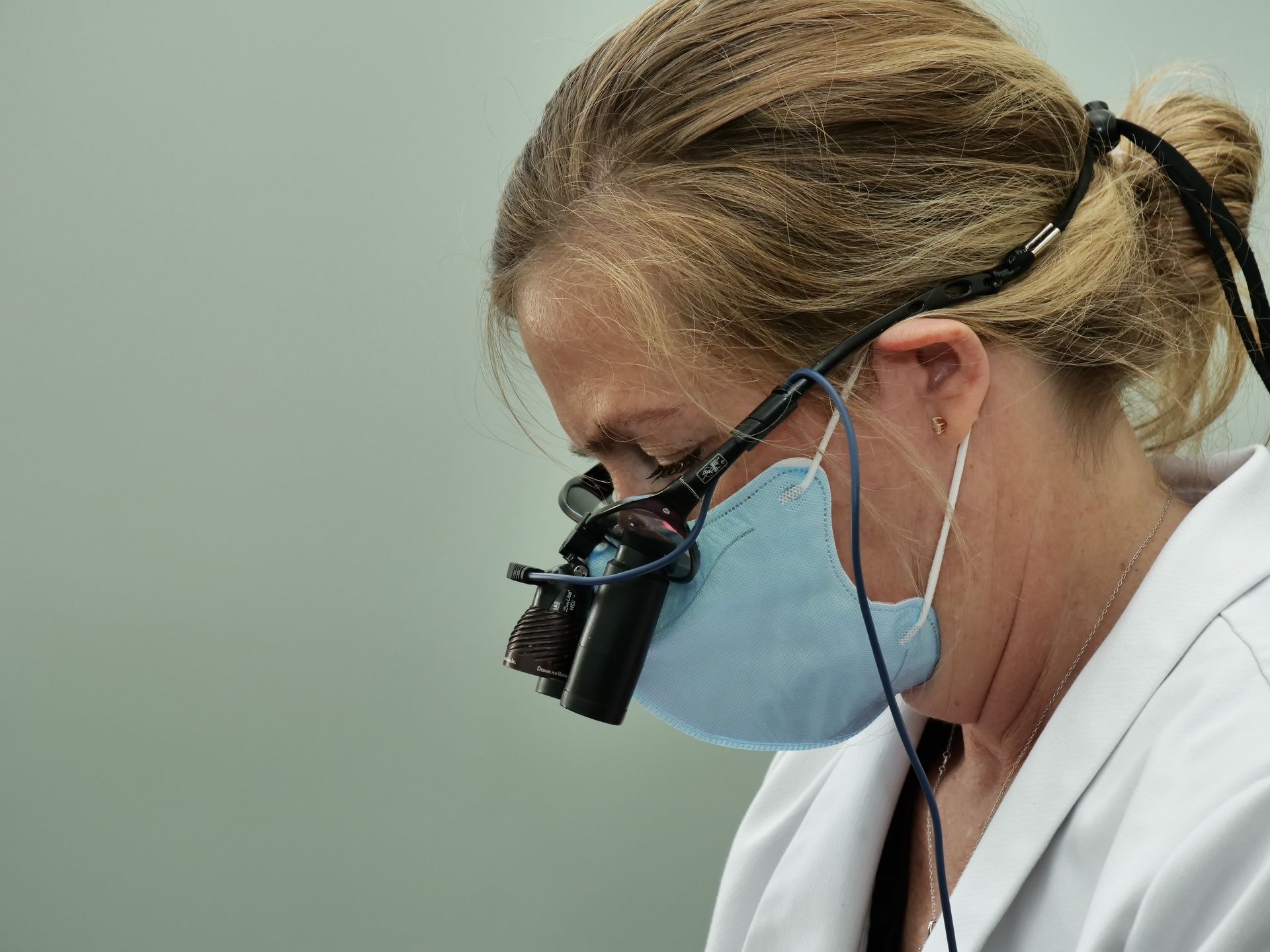 Healthcare worker wearing a blue mask and magnifying visor, leaning over a procedure in a clinical setting