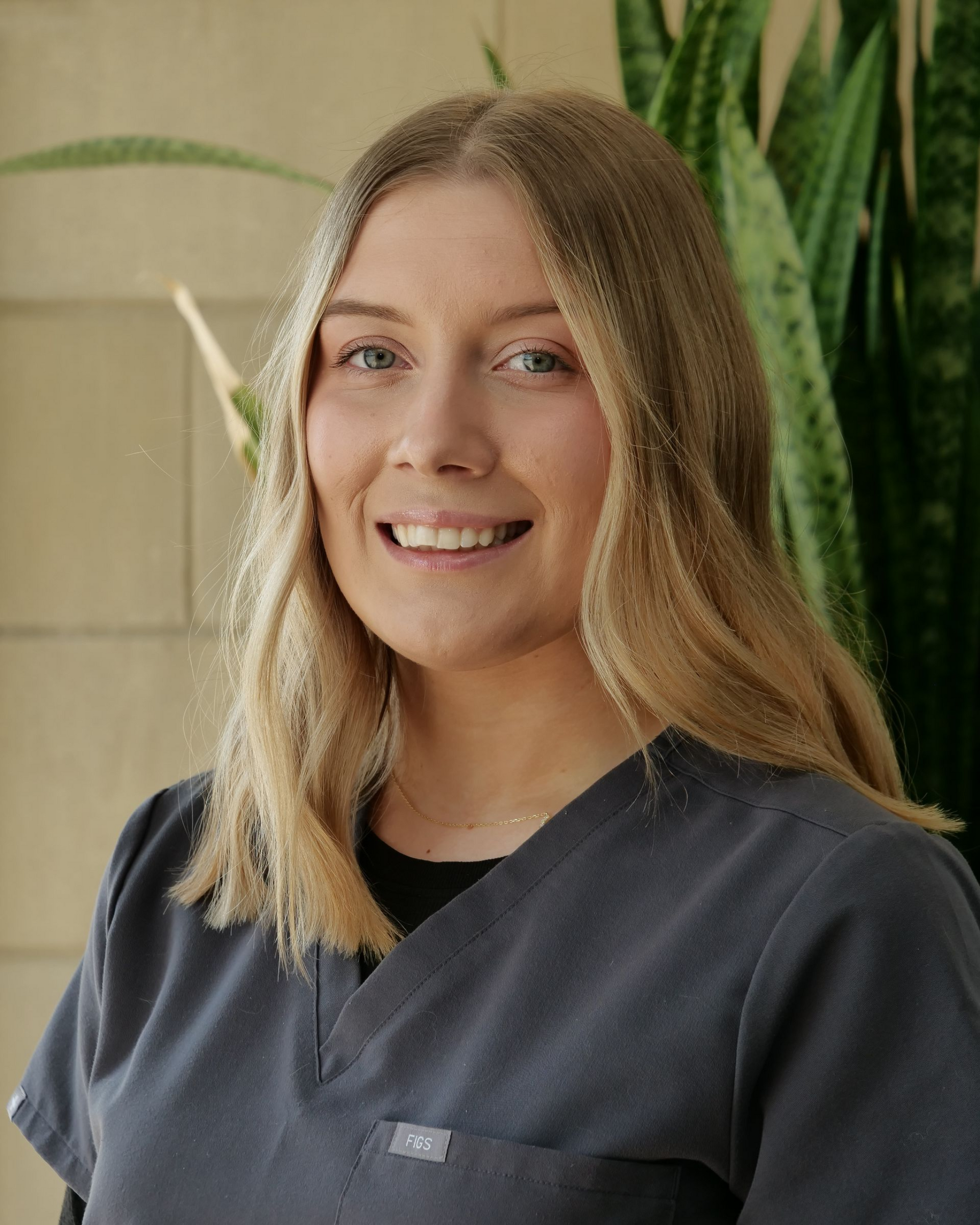Smiling person in a dark shirt standing indoors near a tall green plant.