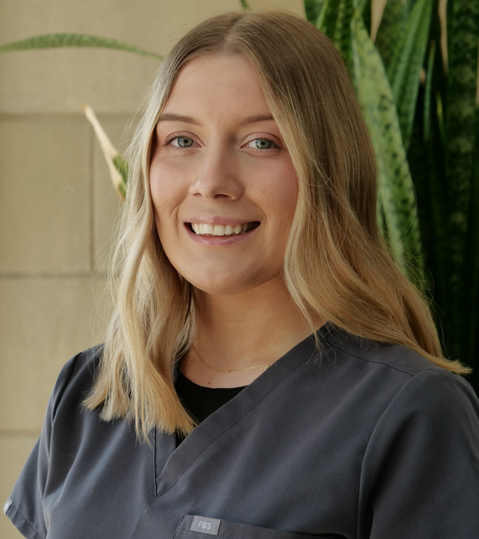 Smiling person in a dark shirt standing indoors near a tall green plant.