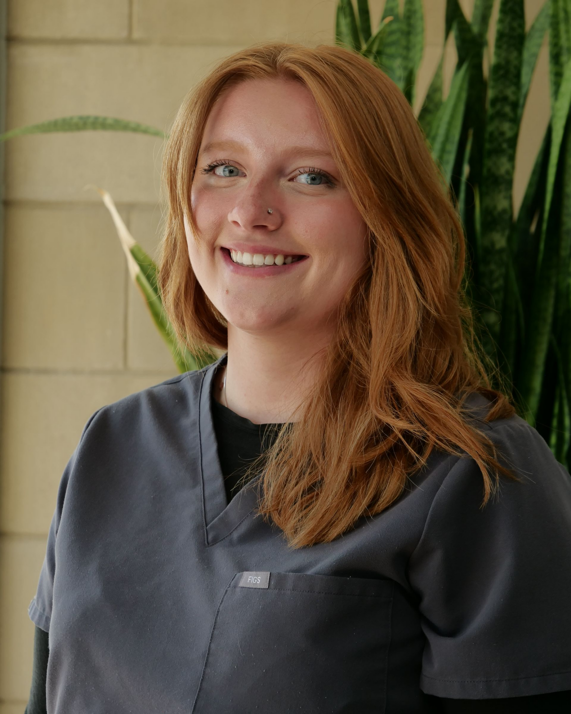 Smiling person in dark gray scrubs standing indoors against a tiled wall with green plants.