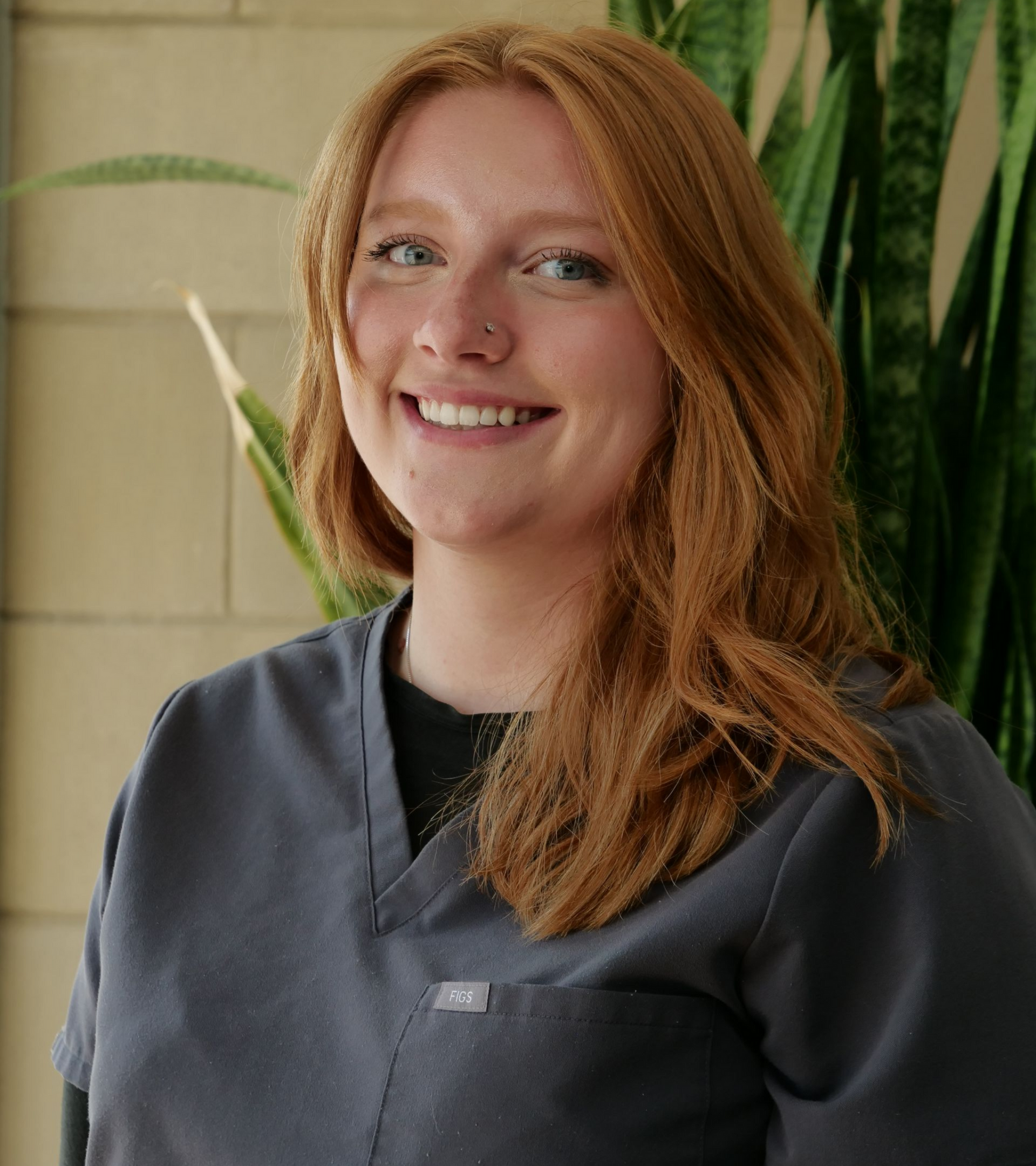 Smiling person in dark gray scrubs standing indoors against a tiled wall with green plants.