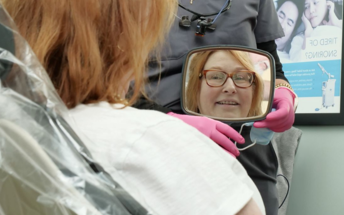 Woman’s face reflected in a handheld mirror during an outdoor event, with a pink-gloved hand holding it.