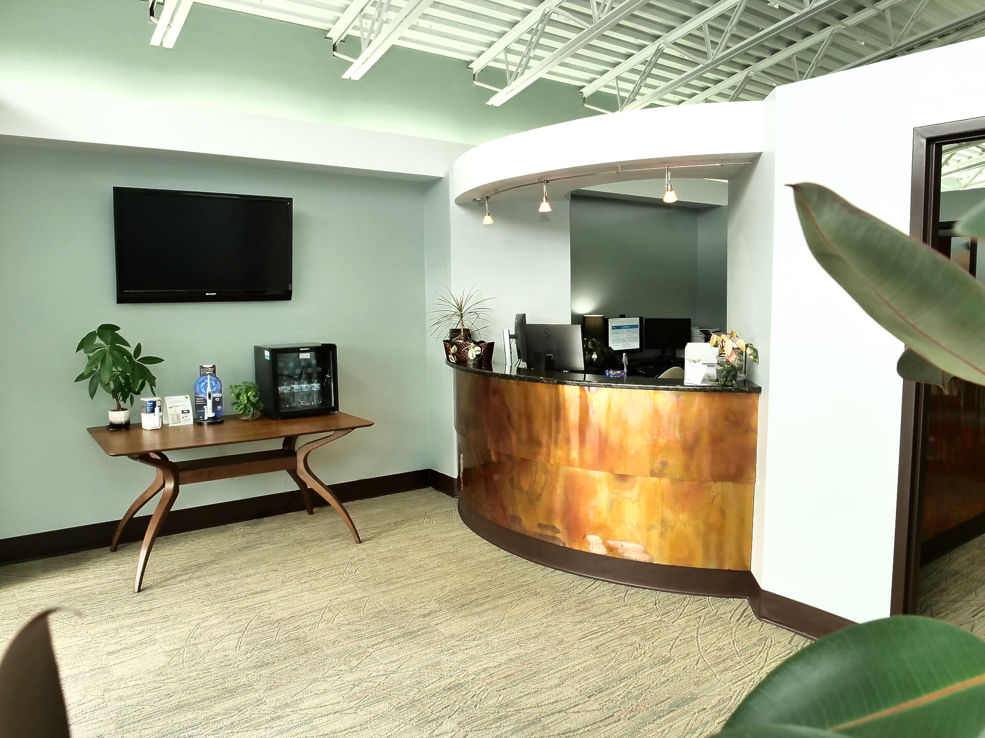 Modern office lobby with curved wooden reception desk, wall TV, and small table with plants and drinks