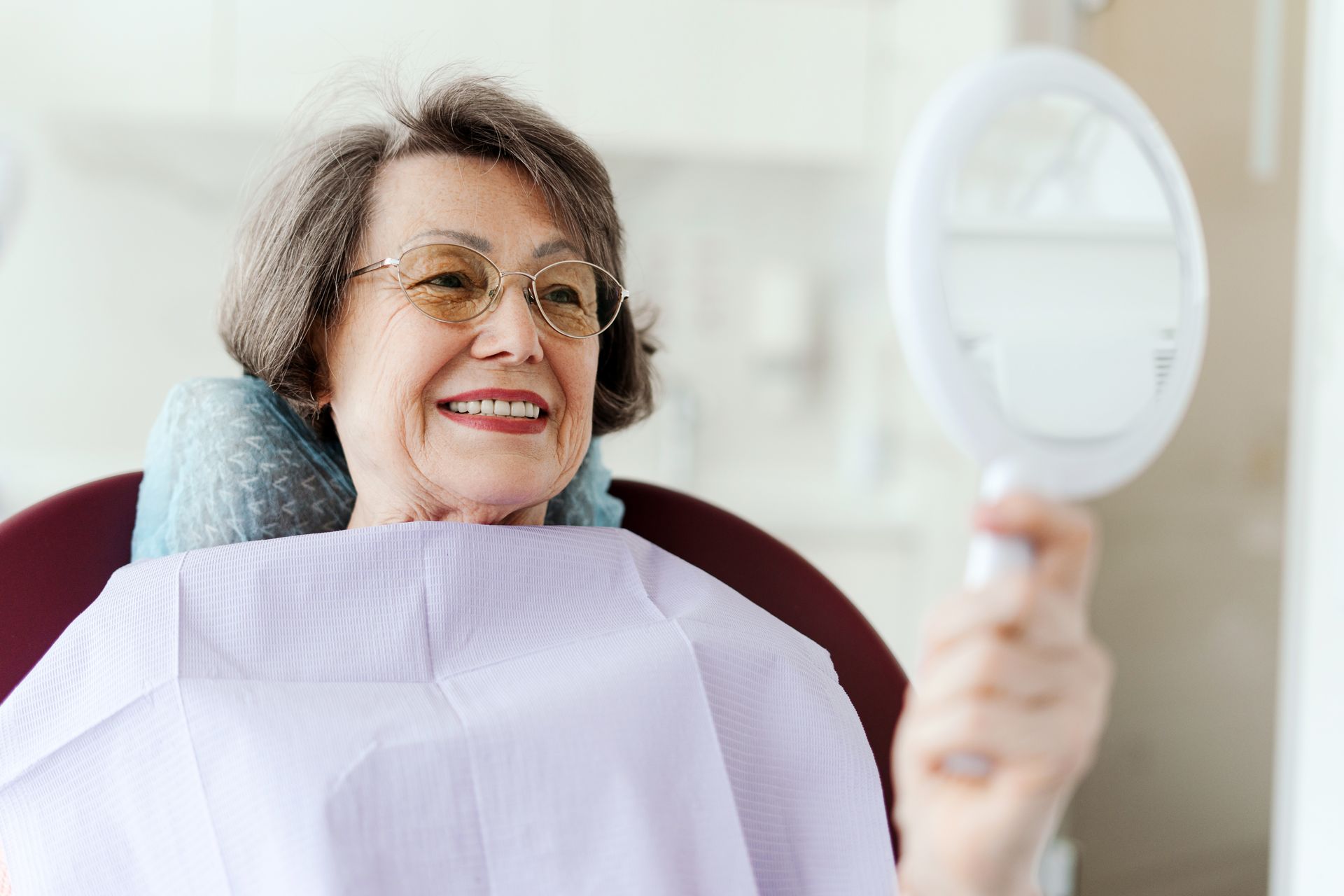 Smiling woman in a dental chair holding a mirror, wearing a bib and looking at her teeth