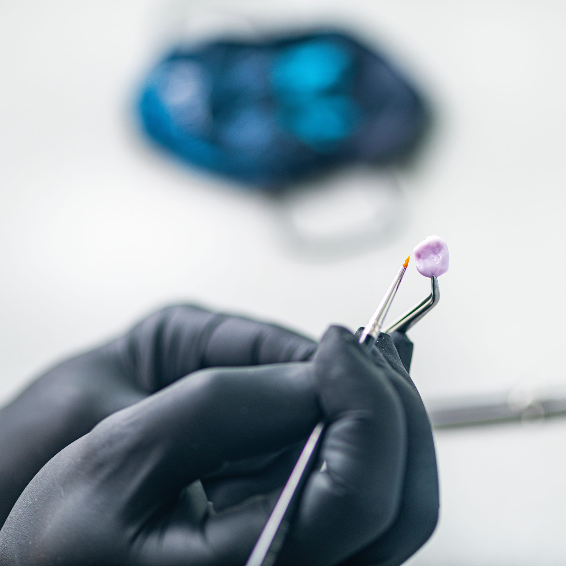 Black-gloved hands holding a dental tool with a purple tip, blurred dental equipment in the background