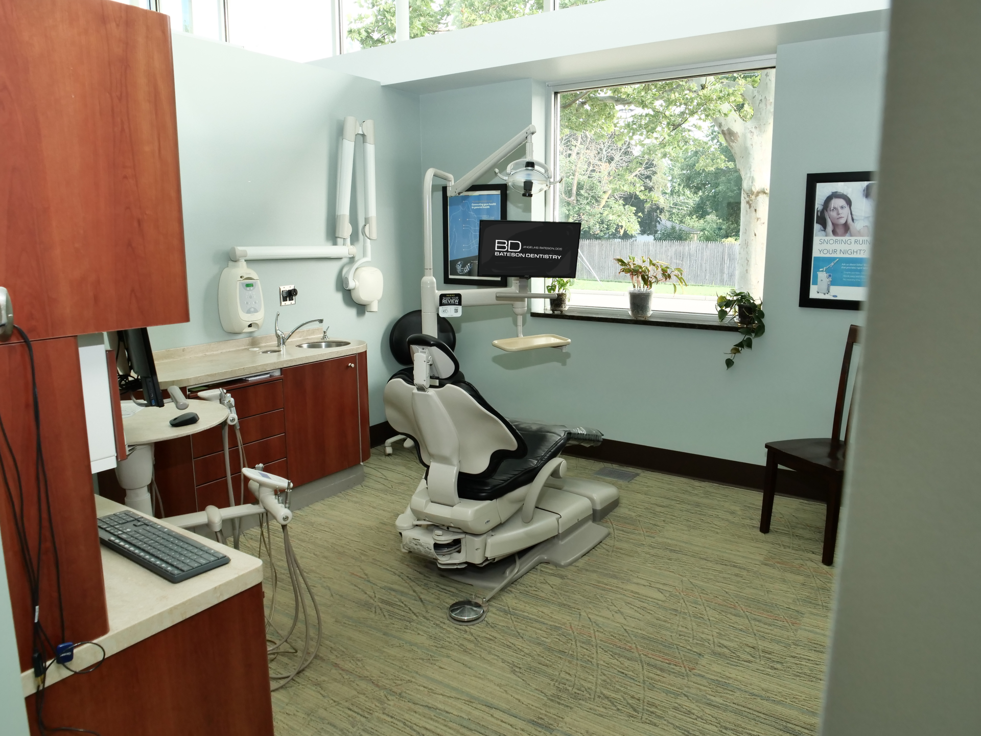 Dental clinic treatment room with chair, monitors, cabinets, and large window.