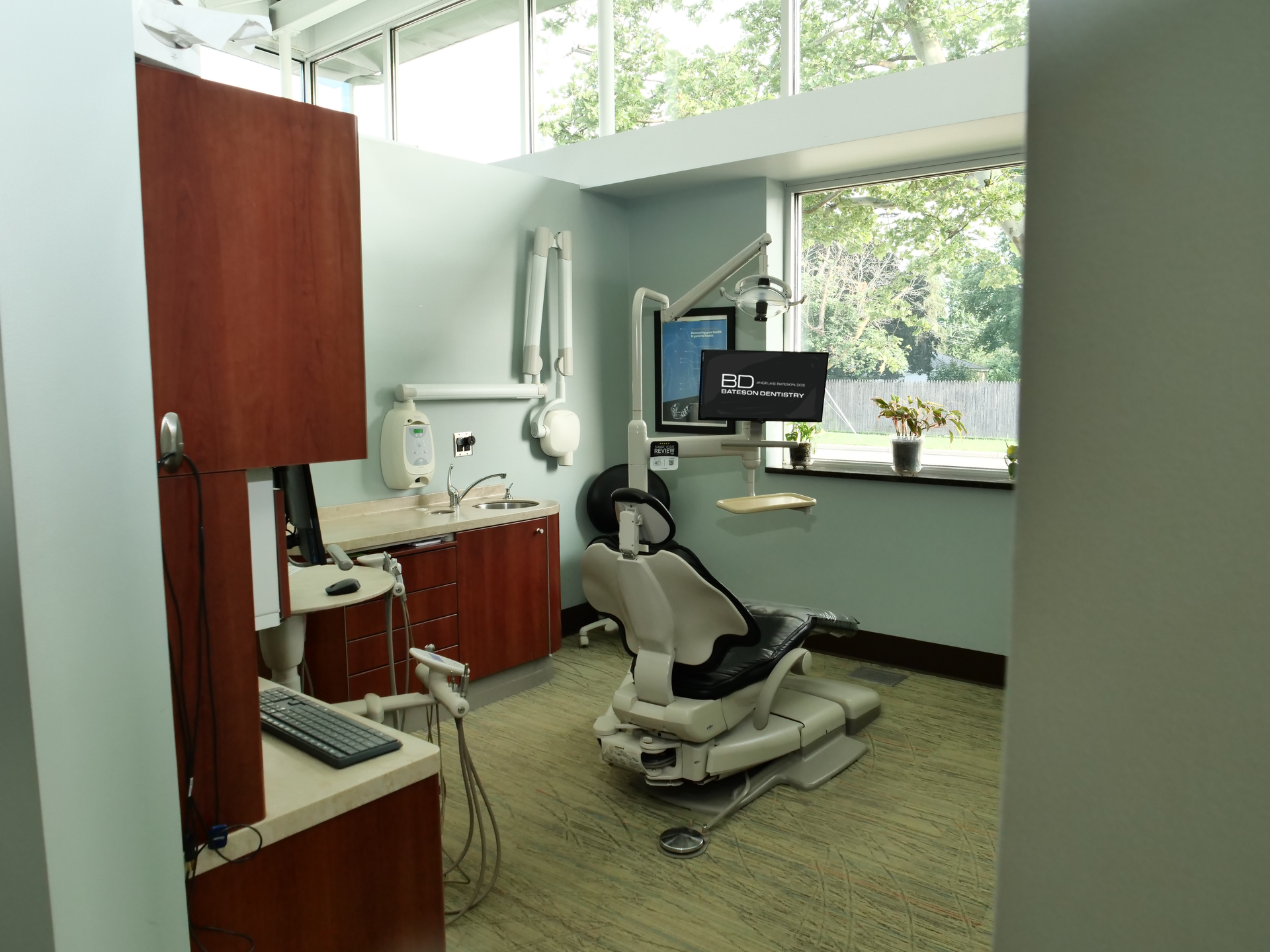 Empty dental exam room with chair, cabinets, monitor, and large windows.