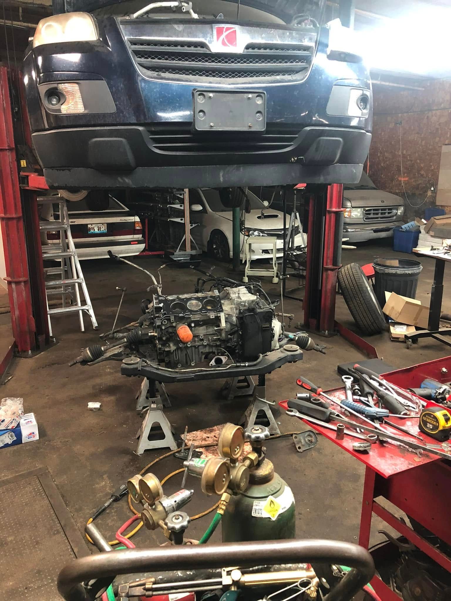 Blue SUV lifted on a shop hoist in a cluttered auto garage, with a removed drivetrain on the floor. | Autotech Service