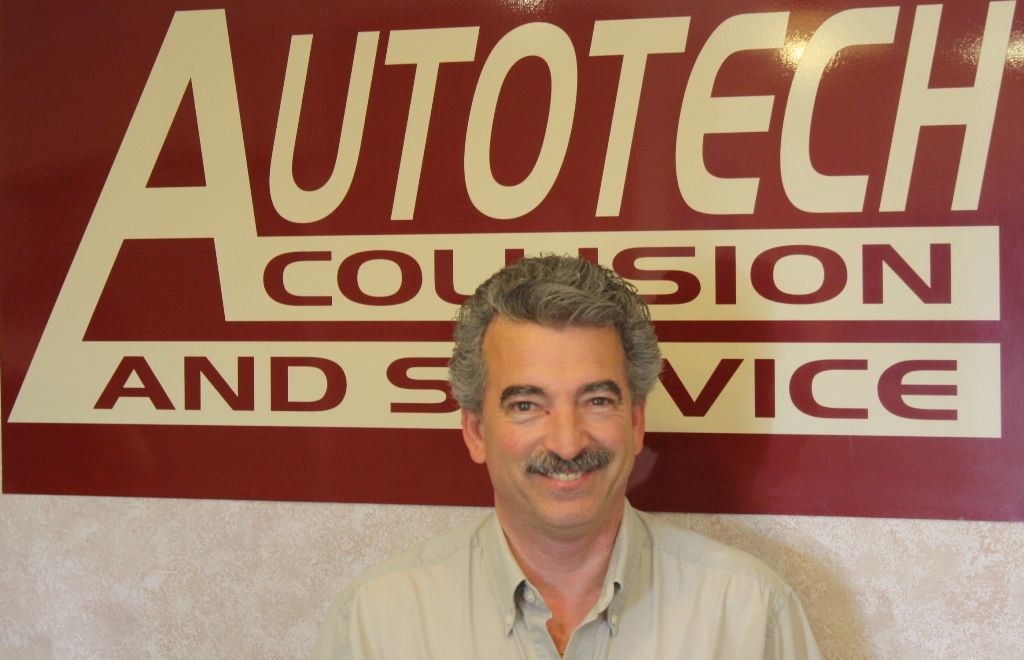 Man standing before a red-and-white “AUTOTECH COLLISION AND SERVICE” sign, smiling | Autotech Service