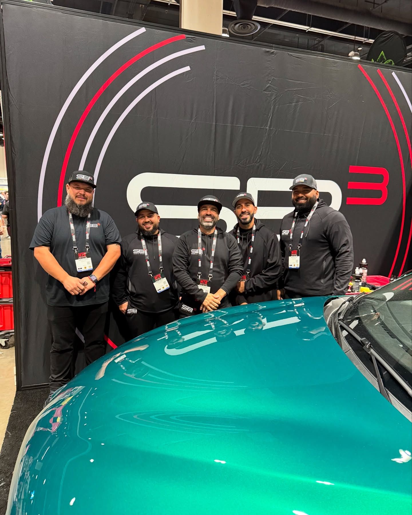 Five men in black shirts pose in front of a turquoise car and a banner with the word 