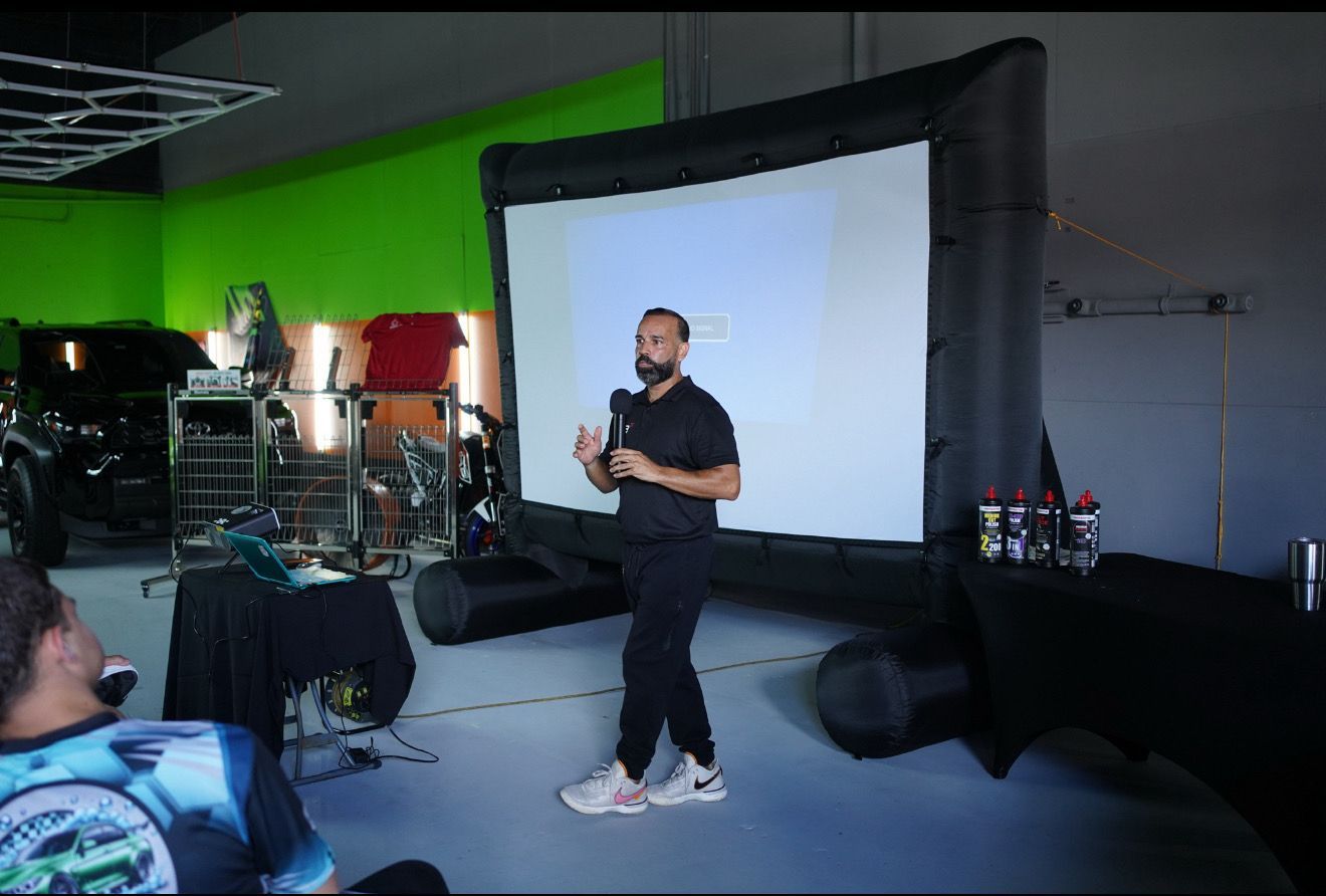 Man demonstrates products to a group, holding two cans, inside a workshop.