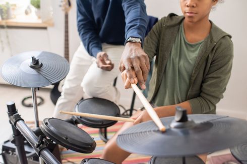 Person teaching drumming to a seated student with an electronic drum kit in a bright room