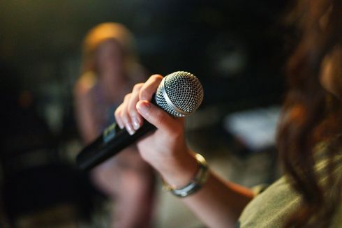 Hand holding a microphone in a dimly lit room, with a blurred person in the background
