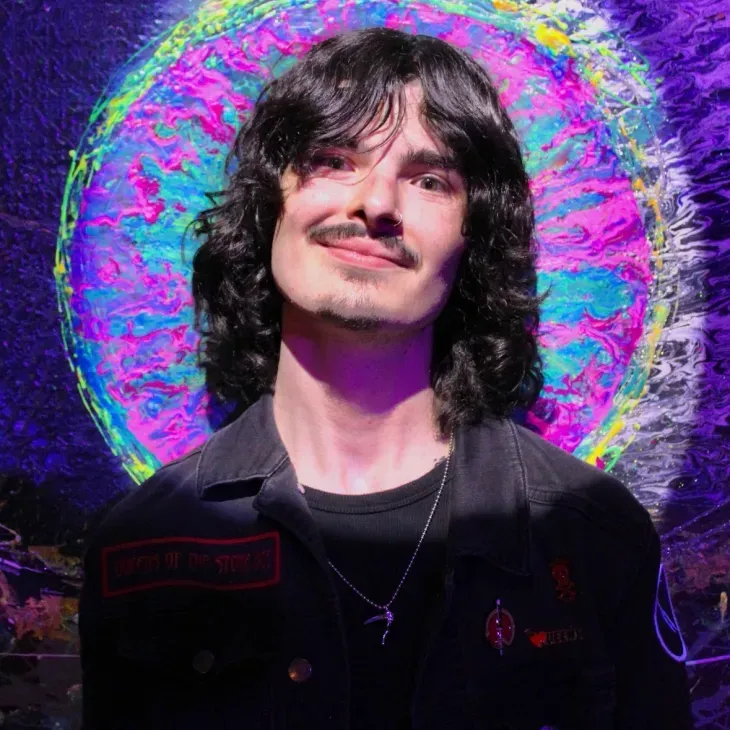 Smiling person with curly hair in a black jacket, standing before a vivid neon halo backdrop.