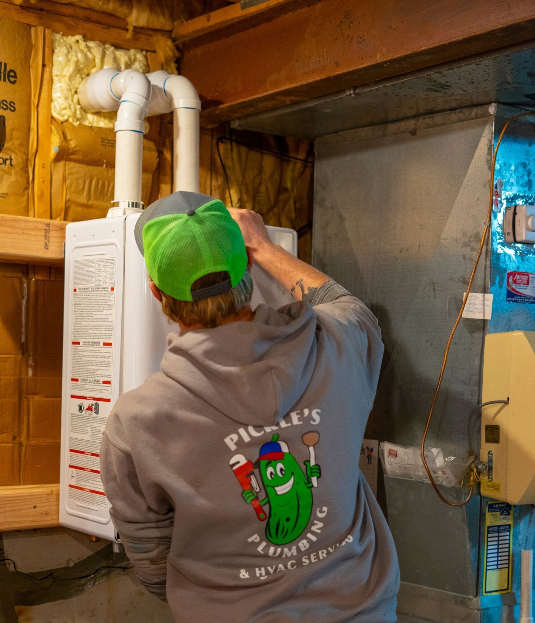 Person inspecting a white water heater and pipes in an unfinished basement, wearing a green cap.