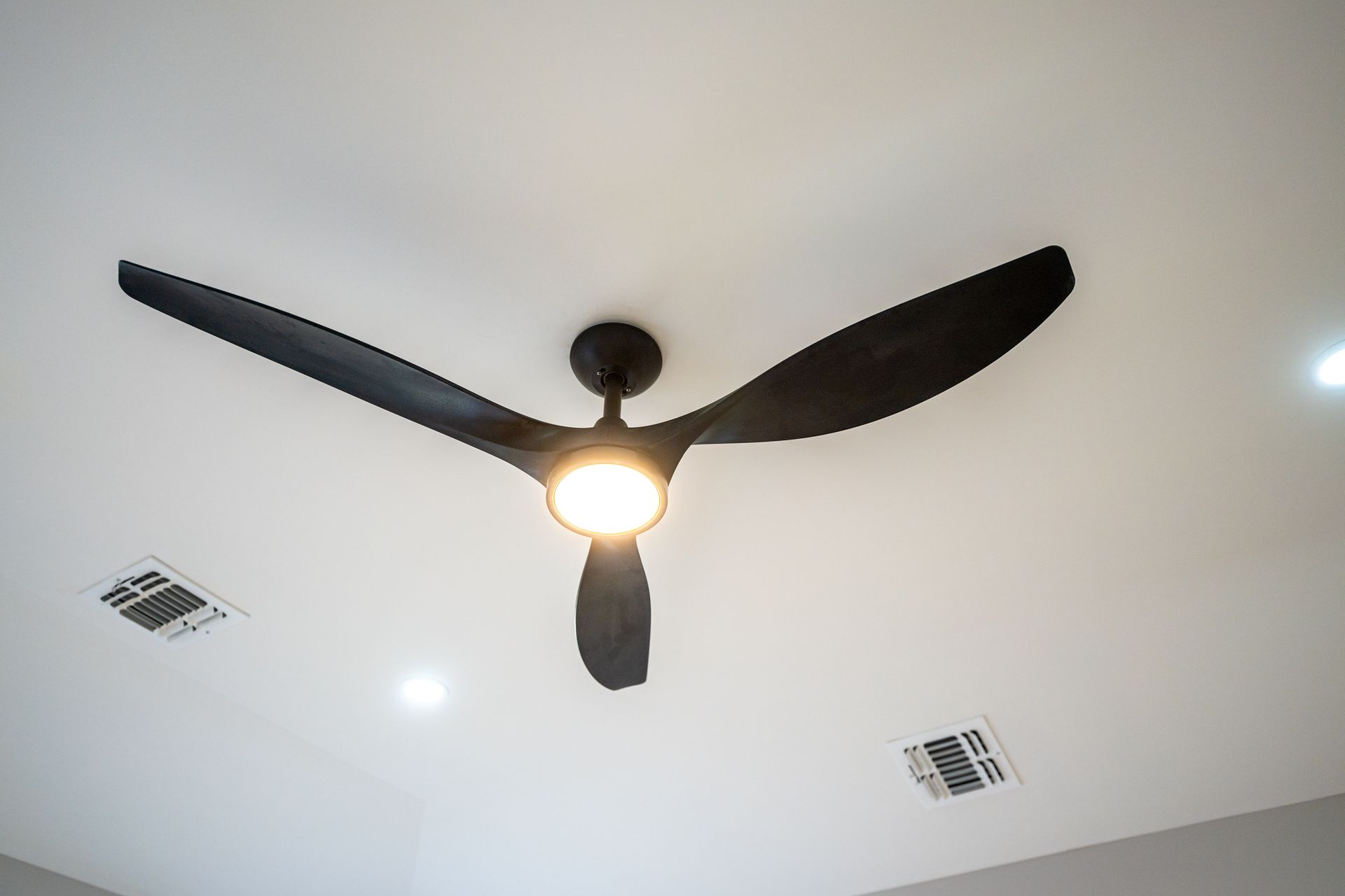 Black, three-blade ceiling fan with integrated light against a white ceiling, two vents visible.