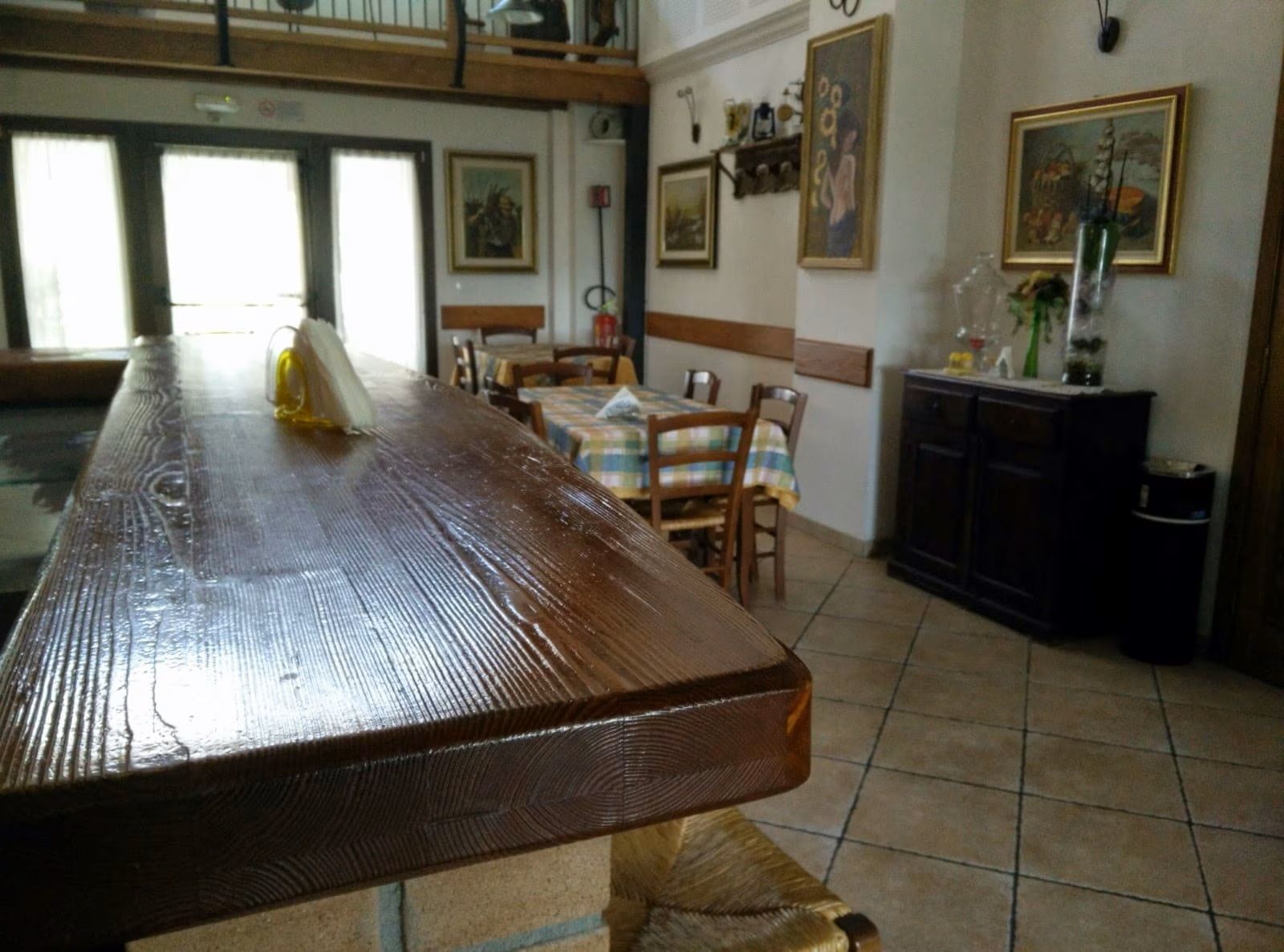 Un lungo tavolo da pranzo in legno in una stanza luminosa con sedie, pavimento piastrellato e quadri appesi alle pareti.