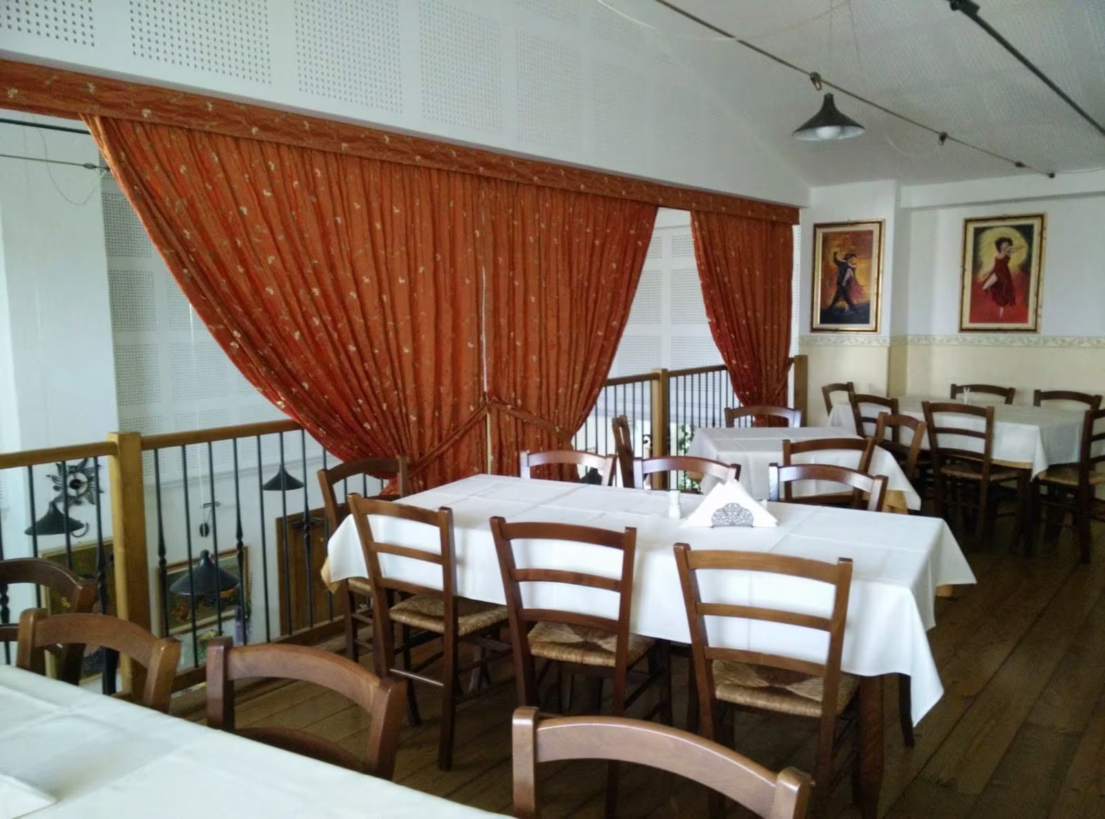 Sala da pranzo con tovaglie bianche, sedie di legno e tende rosse vicino alla ringhiera del balcone.