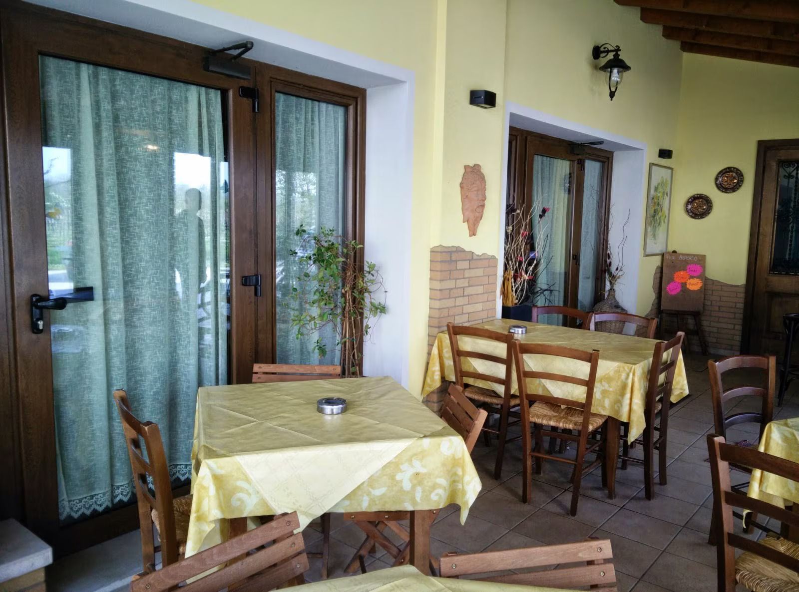 Patio esterno con tavoli e sedie in legno accanto a una porta a vetri e un muro giallo.