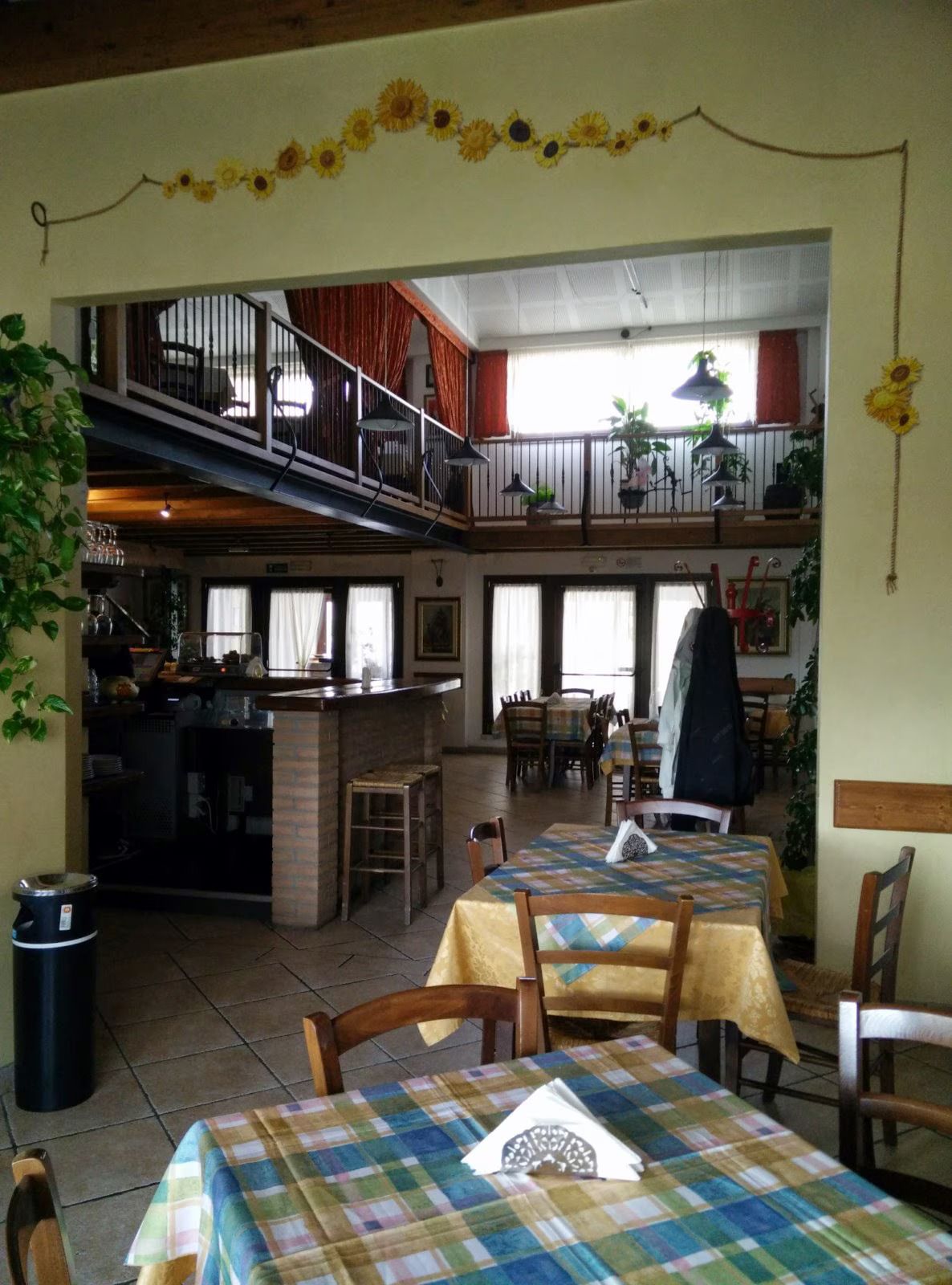Accogliente interno del caffè con tavoli in legno, tovaglie a quadri e una zona con posti a sedere sul balcone al piano superiore.