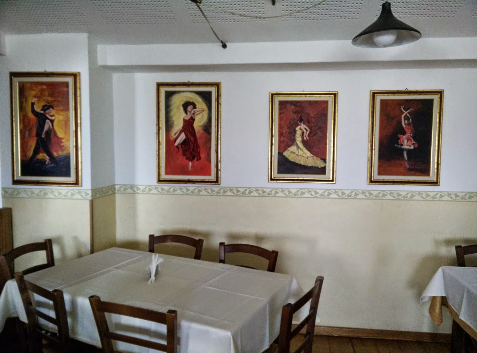 Sala da pranzo con quattro quadri di ballerine incorniciati sopra i tavoli e le sedie di legno, pareti bianche e lampade a sospensione.